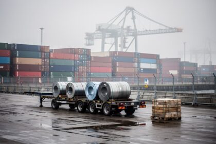 Conteneurs de fret et bobines d'acier empilés sur un quai portuaire industriel européen sous un ciel gris voilé.