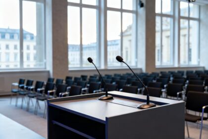 Pupitre équipé de deux micros face à des rangées de chaises vides dans une salle de conférence aux grandes baies vitrées