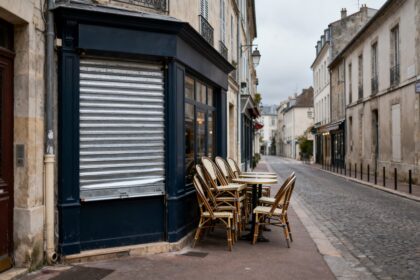 Devanture de commerce fermée par un rideau métallique et chaises de bistrot empilées sur une rue pavée déserte