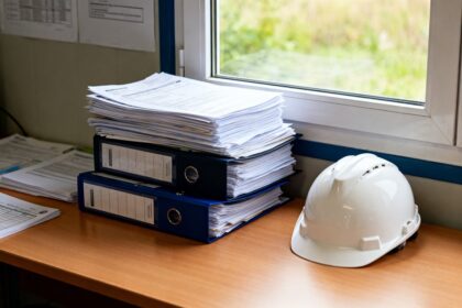 Dossiers administratifs et casque de chantier posés sur un bureau d'entreprise, éclairés par la lumière d'une fenêtre