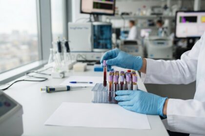 Mains gantées de bleu déposant un tube de sang dans un portoir, sur une paillasse de laboratoire