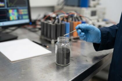 Bocal de poudre grise sur une paillasse, main gantée tenant une spatule, cellules de batterie en arrière-plan