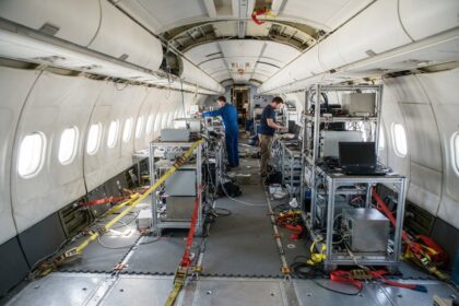 Cabine d'avion sans sièges où des techniciens installent des racks d'expériences sanglés au plancher