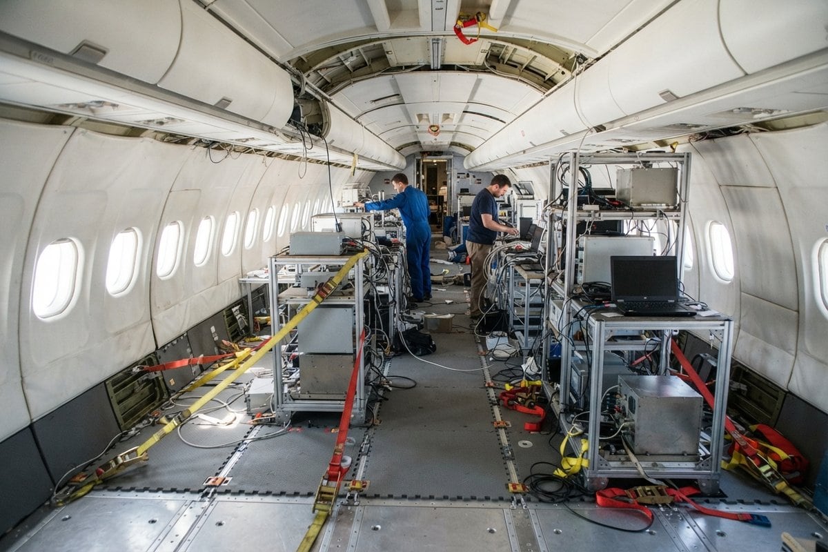 Cabine d'avion sans sièges où des techniciens installent des racks d'expériences sanglés au plancher