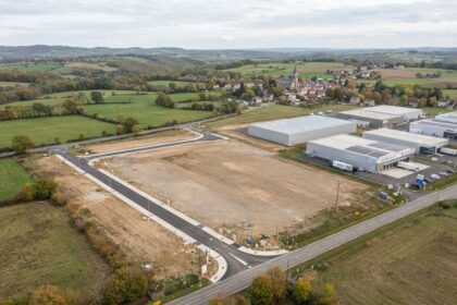 Vue aérienne d'un terrain viabilisé avec voirie neuve, à côté d'entrepôts, devant un village et sa campagne